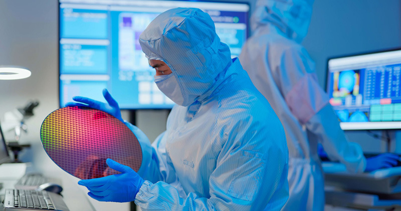 Ein Halbleitertechniker in Reinraum-Schutzkleidung und blauen Handschuhen untersucht einen reflektierenden Siliziumwafer. Digitale Überwachungsbildschirme sind zu sehen, und ein weiterer Techniker arbeitet im Hintergrund.